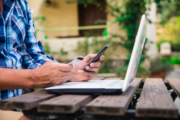 Forty years old caucasian man looking at credit card while working on a laptop computer on a garden terrace during a sunny summer day