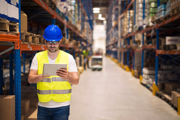 Warehouse worker checking inventory on his tablet while walking in large storage department with shelves and packages in the background.