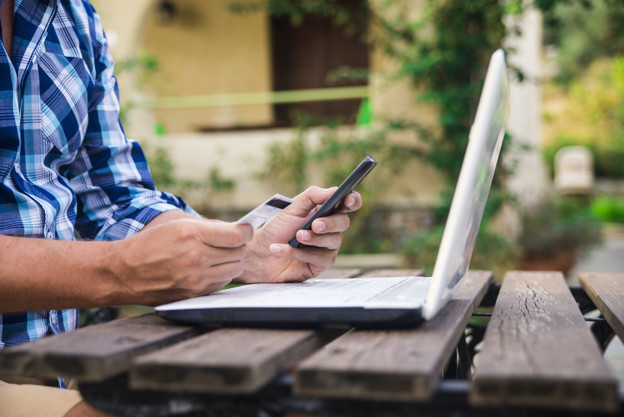 Forty years old caucasian man looking at credit card while working on a laptop computer on a garden terrace during a sunny summer day
